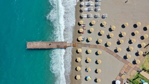 An aerial view of a beach with rows of sun umbrellas and lounge chairs, a wooden pier extending into the water, and a pathway leading to the beach. - ASSOS TROY BEACH HOTEL (Ayvacik)