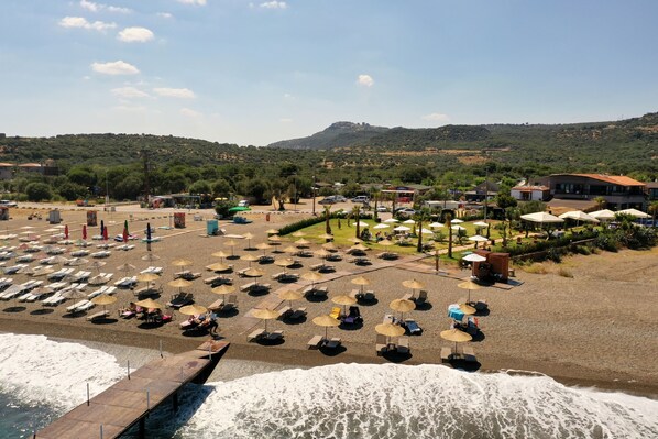 Uma praia com fileiras de espreguiçadeiras e guarda-sóis, um cais que se estende para o mar, e edifícios ao fundo.