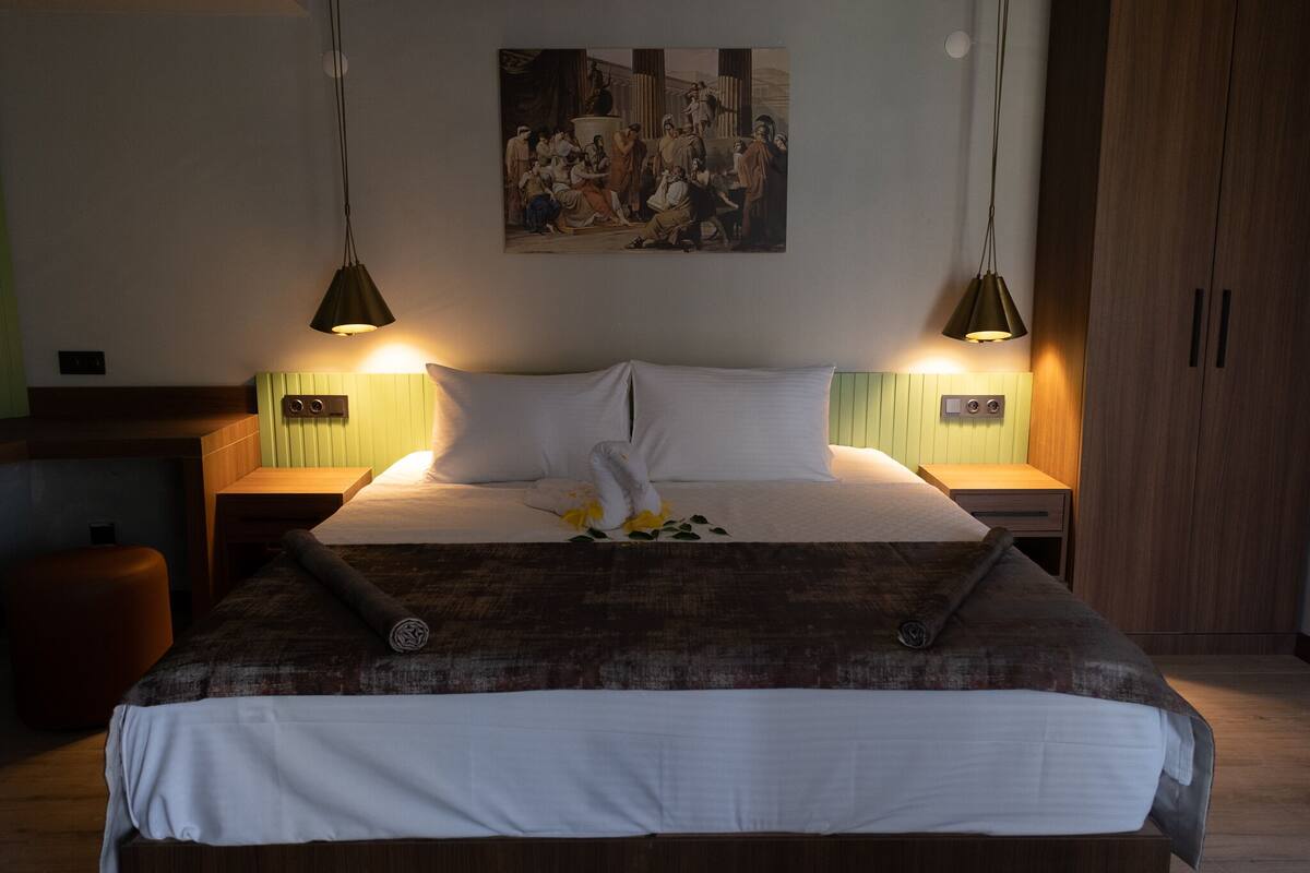 A neatly made bed with white linens, a brown throw blanket, and a decorative pillow. There are two bedside tables with lamps, a wooden wardrobe, and a painting on the wall.