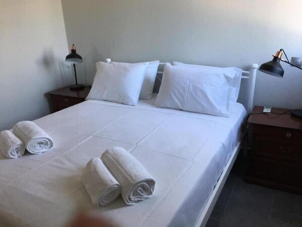 Una cama bien hecha con sábanas blancas y toallas dobladas, una lámpara de noche y una mesita de madera.