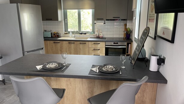 A modern kitchen with a central island, two grey chairs, a built-in oven, and a large window. - Flat - Sleeps 2 - Parking - Patio (Kefalonia)