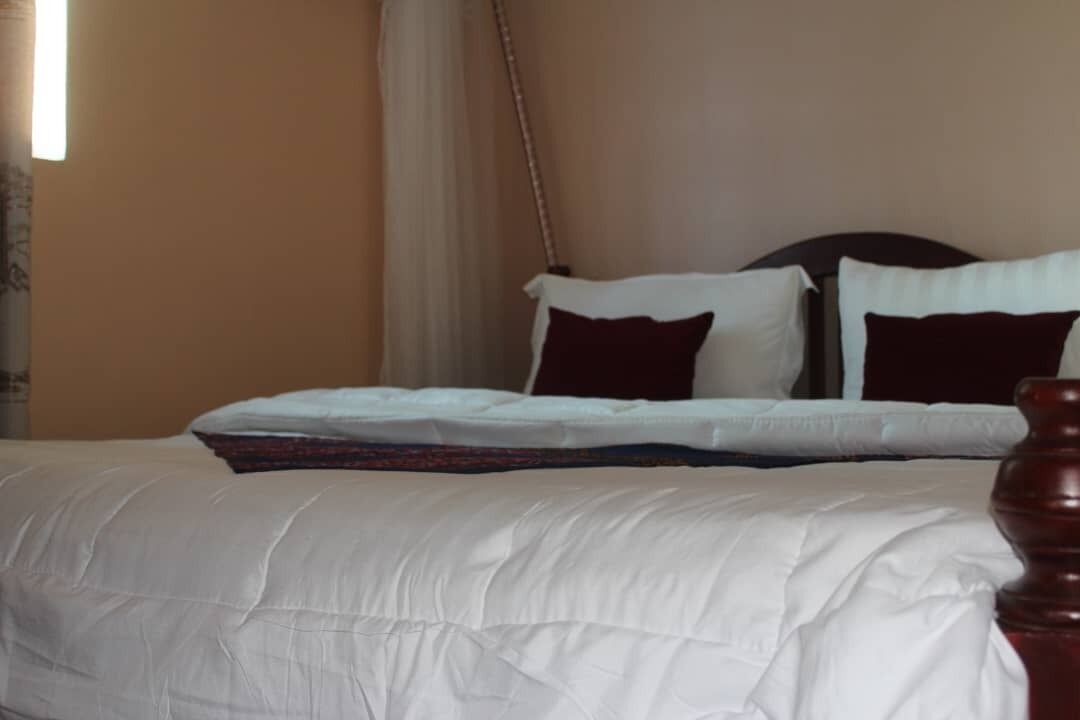 Una cama bien tendida con ropa blanca y almohadas oscuras, un cabecero de madera y una cortina entreabierta.