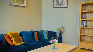 A living room with a dark blue sofa, a white coffee table with a vase of flowers, a bookshelf, and a framed painting on the wall. - The Best Apartment in Canterbury (Canterbury)
