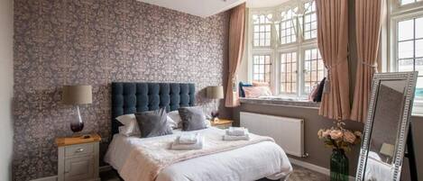 A bedroom with a tufted headboard, a large window with curtains, a bedside table with a lamp, and a mirror.
