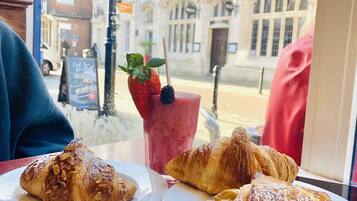 A table with croissants and a coffee cup, viewed through a window overlooking a building with arched windows and a street scene.