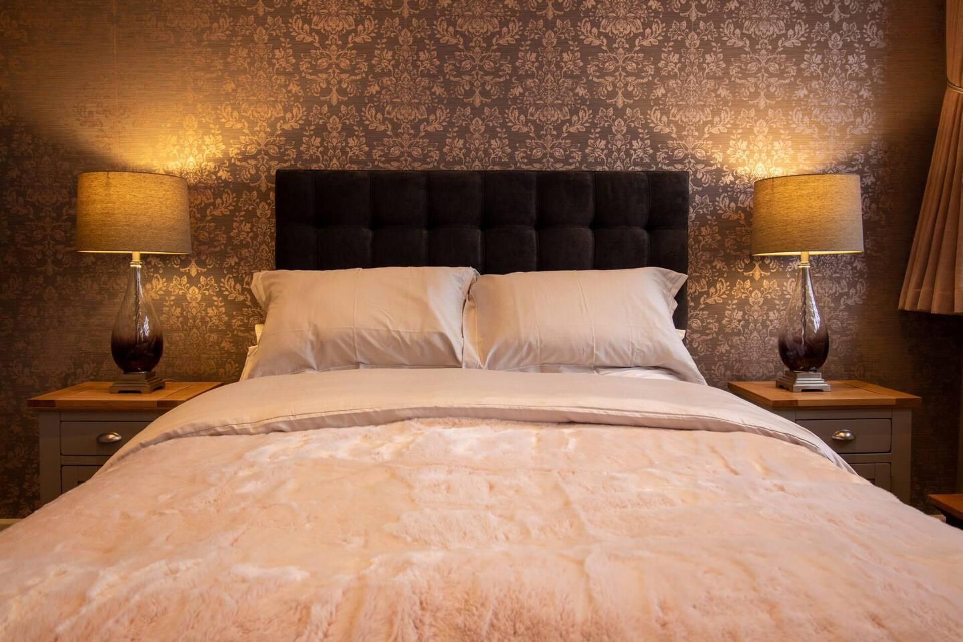 Una cama bien hecha con un edredón blanco mullido, flanqueada por dos mesitas de noche con lámparas, sobre un fondo de papel tapiz estampado.