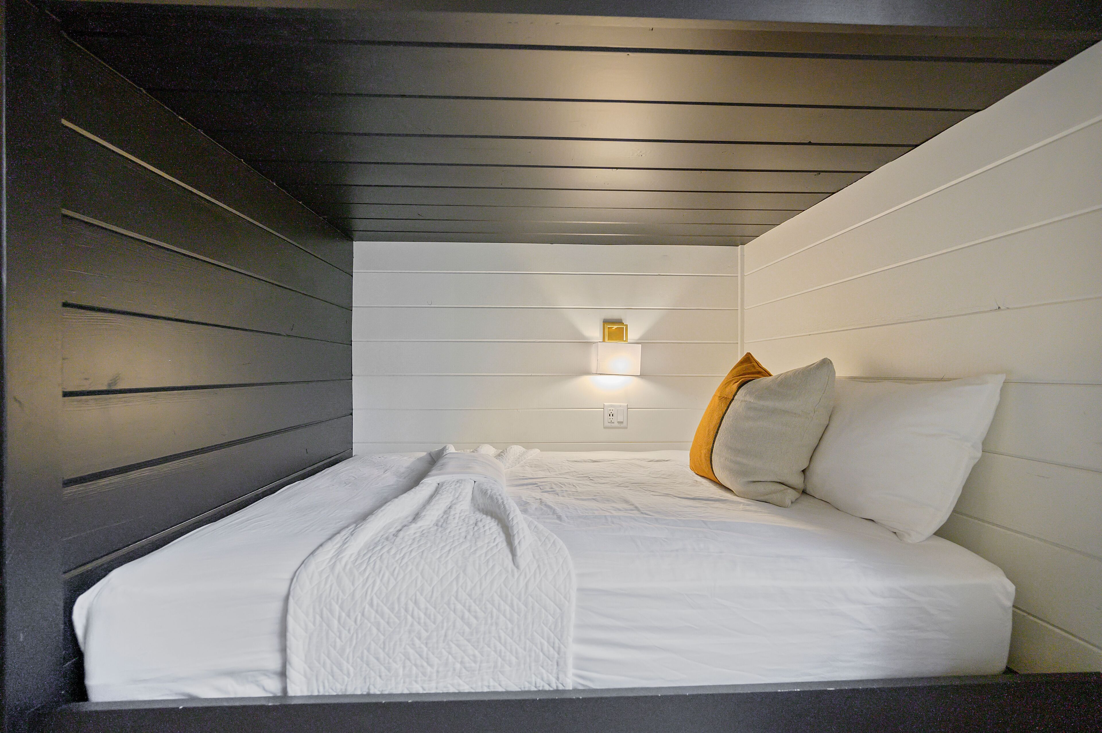 A neatly made bed with white linens and a quilted blanket, a bedside lamp, and a wall-mounted light fixture.