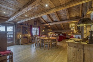 A rustic wooden interior with a wooden dining table and chairs, a kitchen area with a stove, and a large window with curtains. - La Grange Paccaly, 4* Appartment, Heart Village (La Clusaz)
