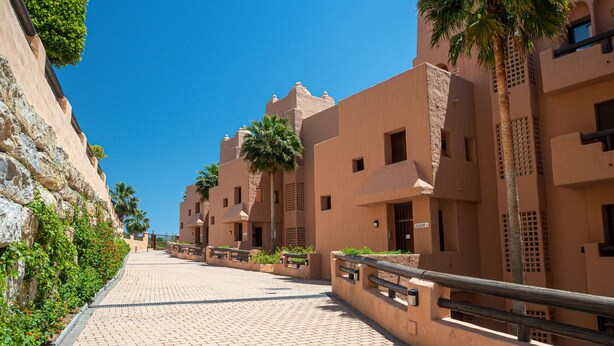 A pathway lined with traditional buildings, palm trees, and a clear blue sky. - Coto Real 2311 Sea View (Manilva)