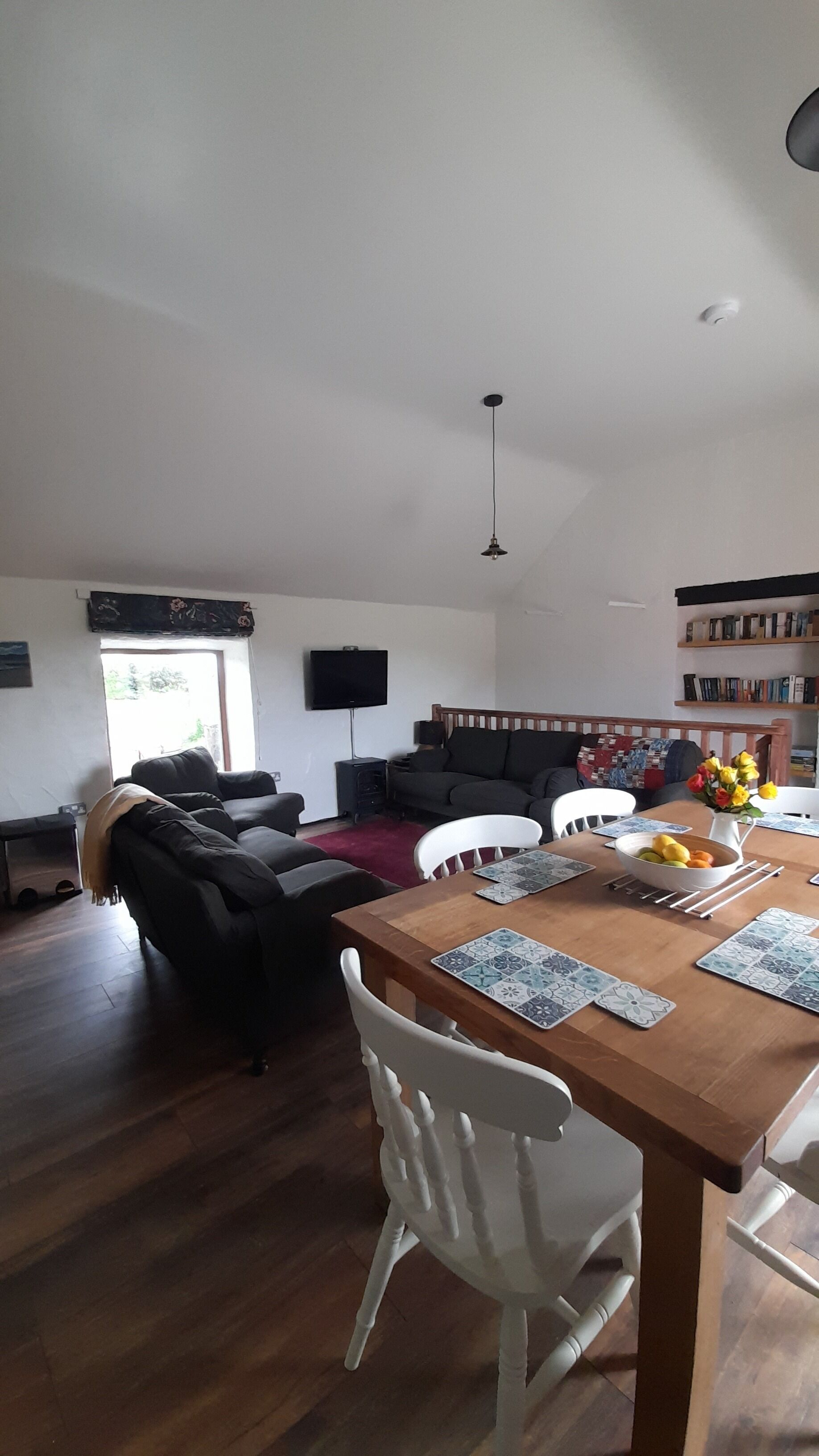 A living room with a wooden dining table, white chairs, a black sofa, and a television on the wall.