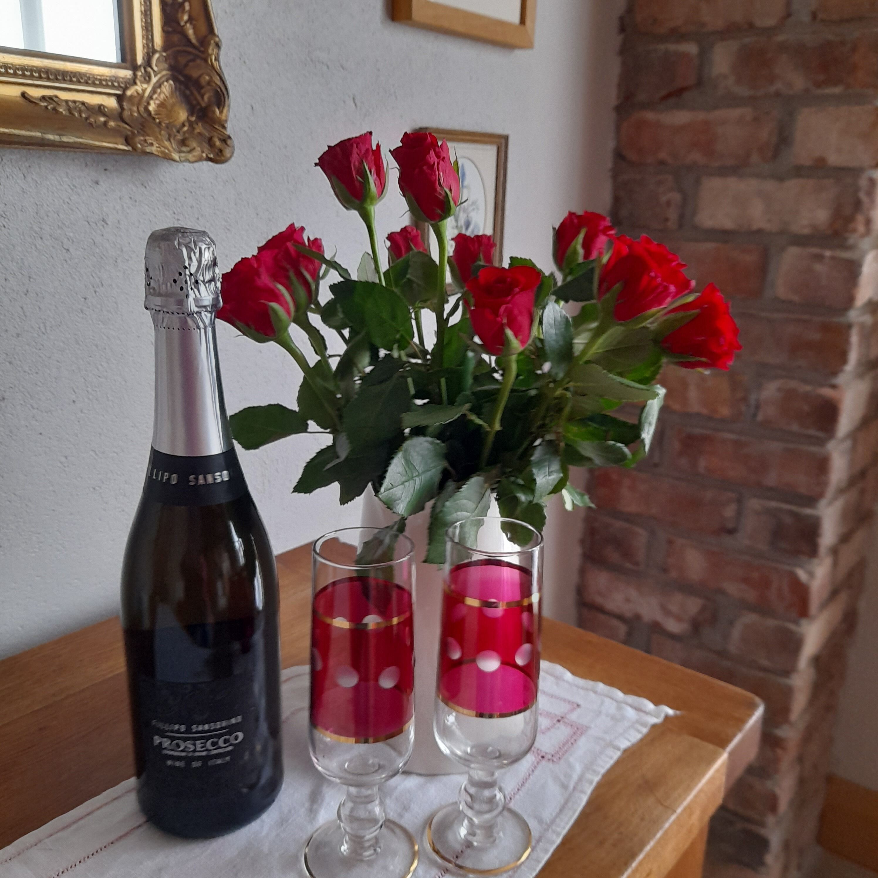 Een fles prosecco, twee glaasjes met rode vloeistof, en een vaas met rode rozen op een houten tafel.