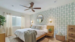Una camera da letto con un letto, una ventola a soffitto, un comodino con una lampada, uno specchio, una finestra con tende e una parete decorata con un motivo a foglia.