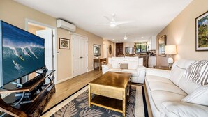 A living room with a TV, white sofa, wooden coffee table, and a kitchen in the background.
