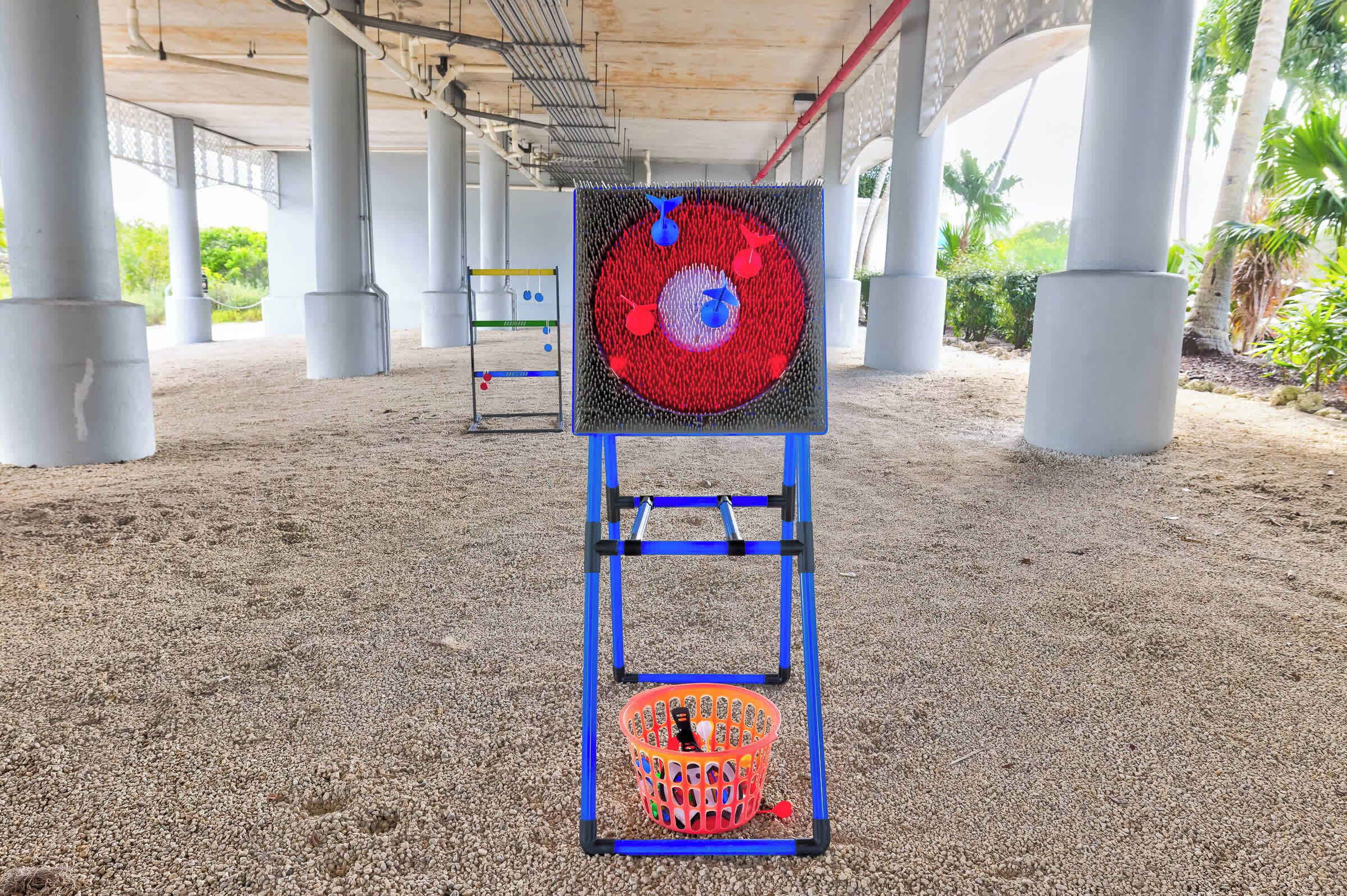 A target with a red bullseye and a basket below it, set up on a sandy ground under a structure with columns.