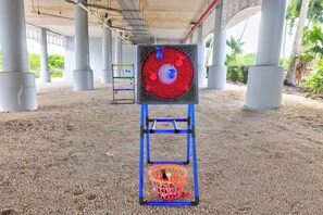 Une cible avec un cercle rouge au centre et un panier en dessous, placée sur un sol sablonneux, sous une structure soutenue par des colonnes.