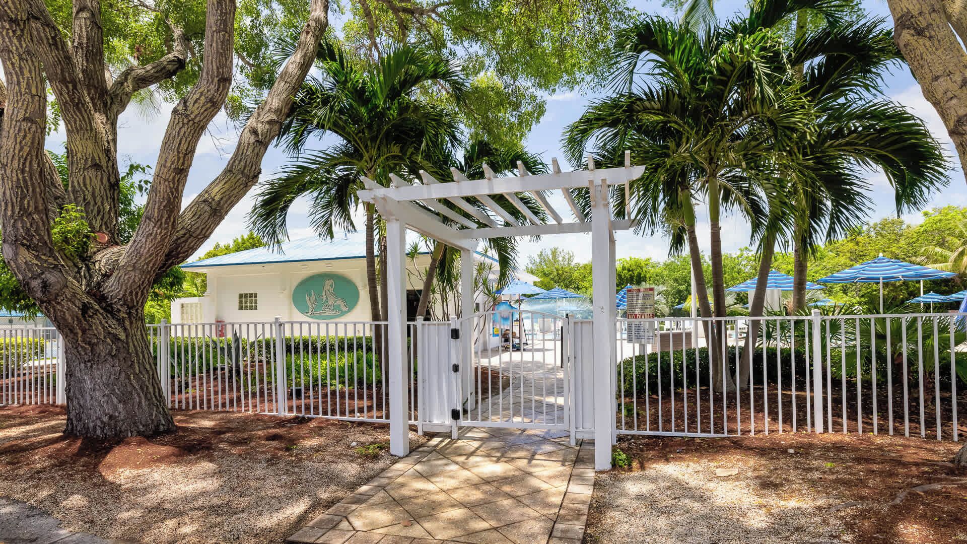 A gated entrance with a pergola, palm trees, and a building with a green logo.