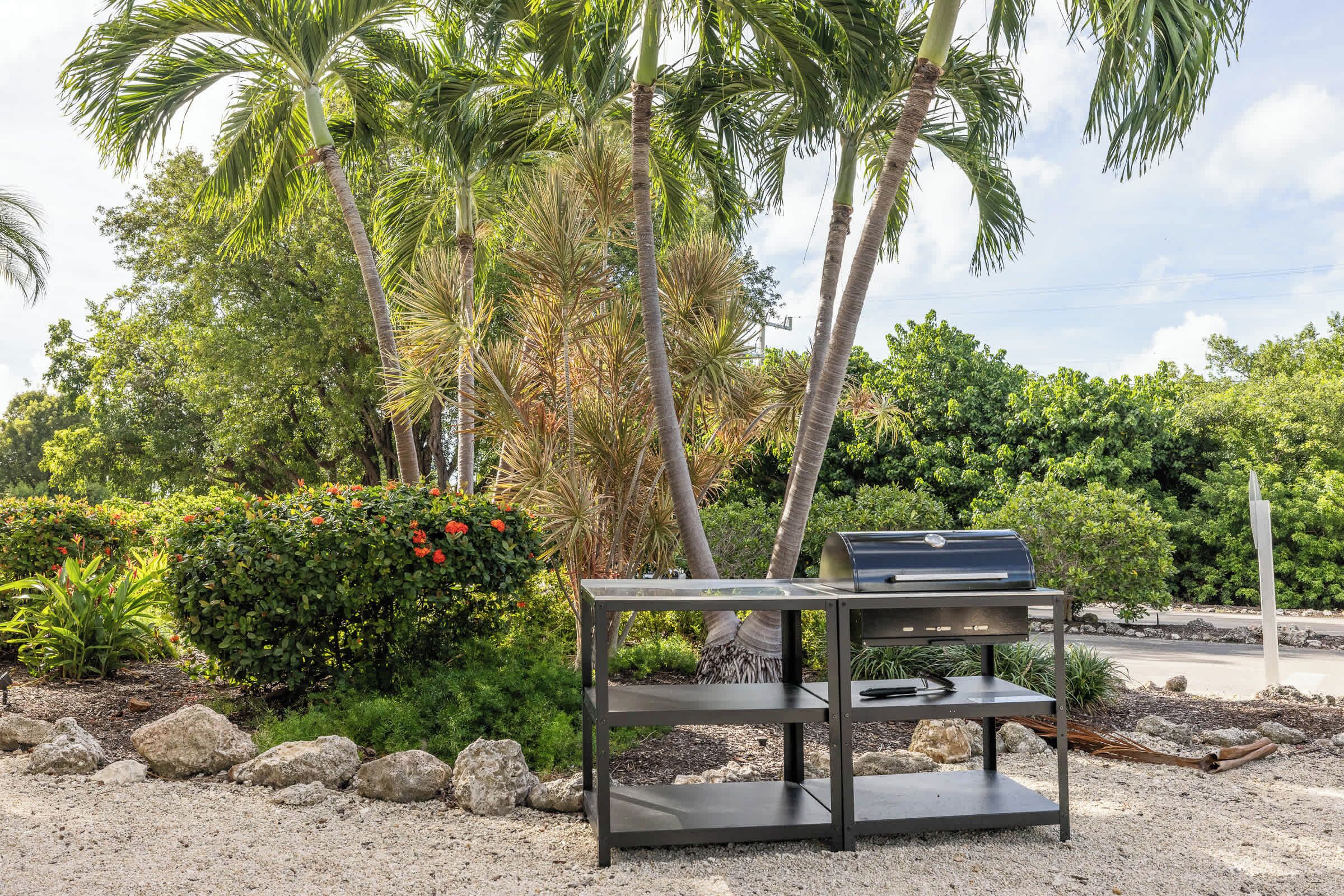 Una parrilla para barbacoa sobre un soporte metálico en un jardín con palmeras y flores rojas.