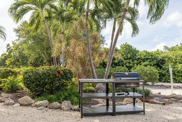 Un barbecue sur un support métallique, dans un jardin agr émenté de palmiers et de fleurs rouges.
