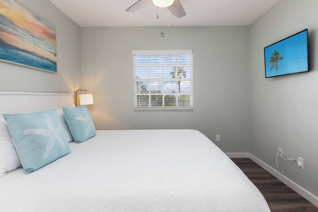 A bedroom with a bed, a ceiling fan, a window with blinds, and a wall-mounted TV.