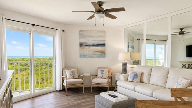 A spacious living room with a large window, a ceiling fan, a sofa, two armchairs, a coffee table, and a side table with lamps.