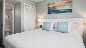 A bedroom with a large bed, white headboard, and a painting of a sunset.