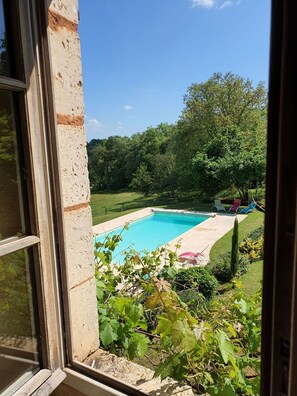 Une vue sur une piscine entourée d’un jardin luxuriant et d’arbres, vue à travers une fenêtre.