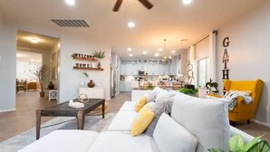 A modern living room with a white sofa, yellow cushions, a wooden coffee table, and a yellow armchair. There is a kitchen in the background with white cabinets and a dining area.