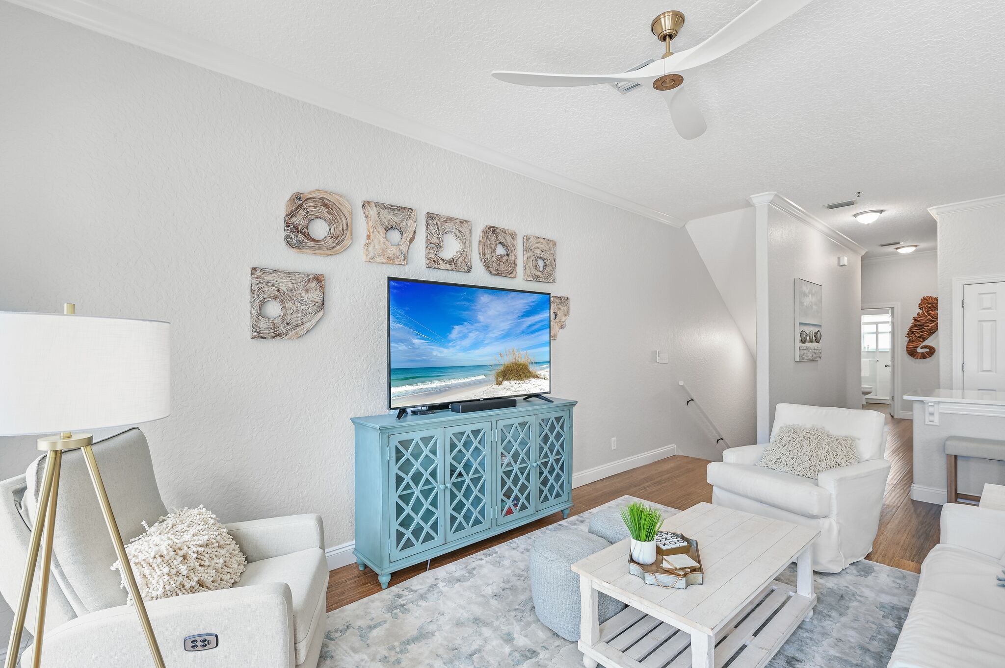 A living room with a TV, white furniture, a ceiling fan, and decorative wall art.