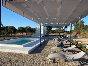 Uma área de piscina com espreguiçadeiras sob um toldo, rodeada por cascalho e árvores.