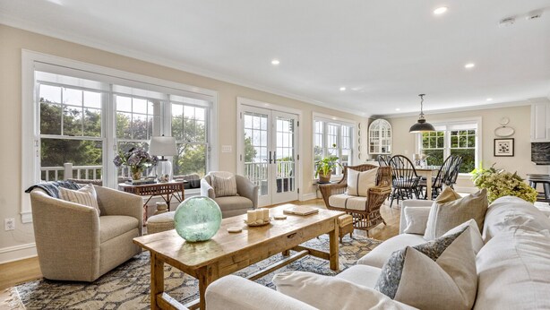 A spacious living room with a large window, a dining area with wicker furniture, and a kitchen visible through an open door. - Beautiful Southampton Home with breathtaking waterfront views, front porch,... (Southampton)