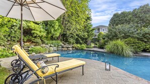 A pool area with two sun loungers, a patio umbrella, and a stone wall with greenery. - Beautiful Southampton Home with breathtaking waterfront views, front porch,... (Southampton)