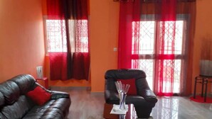 Ein Wohnzimmer mit einem schwarzen Ledersofa, einem schwarzen Sessel und einem Glastisch. Es gibt eine blaue Pendelleuchte, rote Vorhänge und eine Klimaanlage an der Wand.