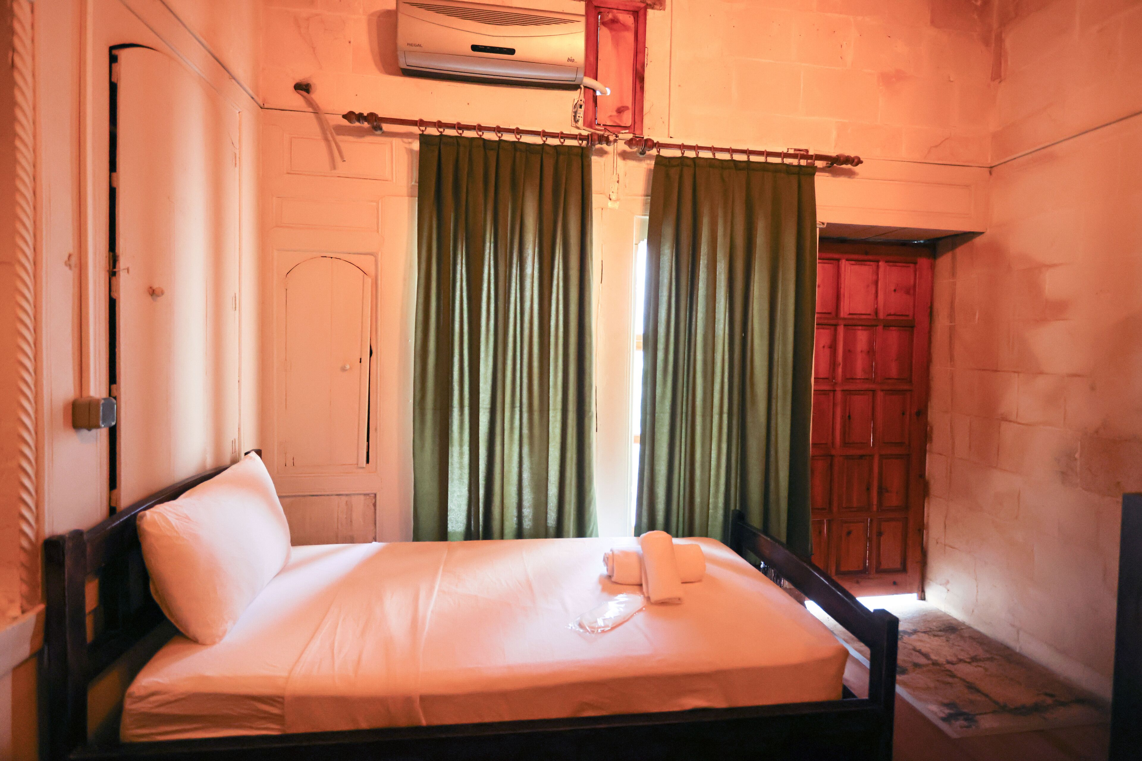 Una cama individual con estructura de madera, colchón blanco y almohada. Se observa una ventana con cortinas verdes, una puerta de madera y un equipo de aire acondicionado fijado a la pared.