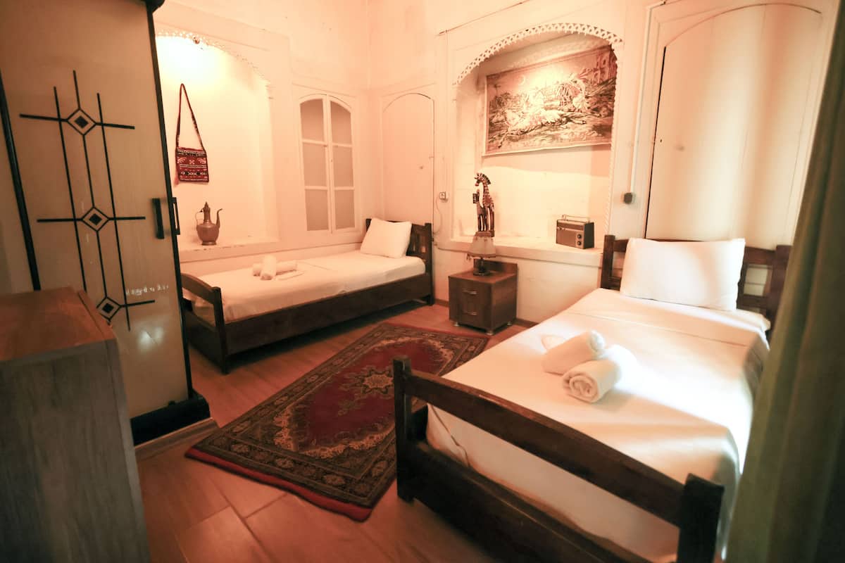 A hotel room with two single beds, a wooden wardrobe, a nightstand, a lamp, and a decorative wall piece.