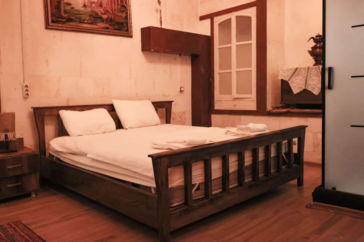 A wooden bed with white linens and a headboard, a nightstand with a lamp, and a window with a wooden frame.