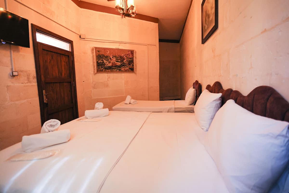 A hotel room with two beds, a wooden door, a painting on the wall, and a TV mounted on the wall.