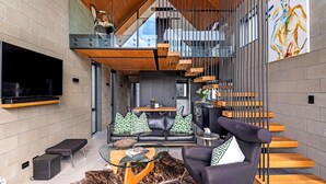Ein modernes Wohnzimmer mit einem großen Flachbildfernseher, einem Glastisch, einer Couch mit gemusterten Kissen und einer Treppe mit Holzstufen.