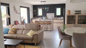A modern living room with a sofa, coffee table, and dining area. There is a kitchen with a dining table and chairs. The room has large windows and a ceiling light fixture. - large pool and jacuzzi villa (Lunel)