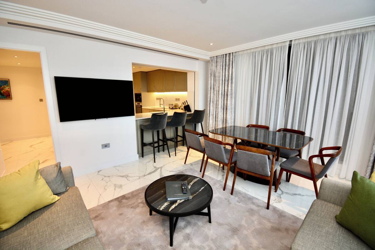 Een moderne woonkamer met een flatscreen-tv, een eethoek met een glazen tafel en stoelen, en een keuken die door een open doorgang zichtbaar is.