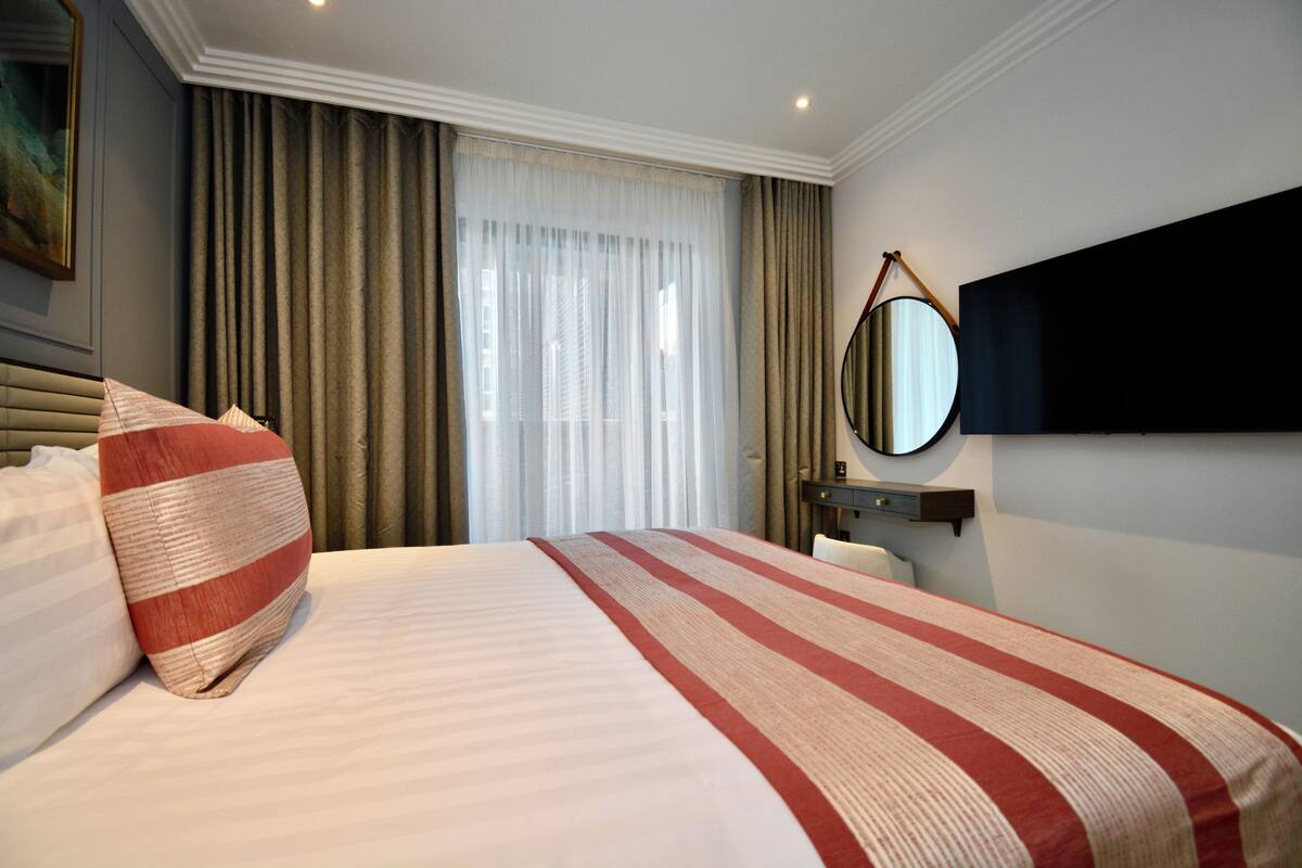 Hotelkamer met een groot bed, nachtkastje, spiegel en uitzicht op het buitenleven door dunne gordijnen.
