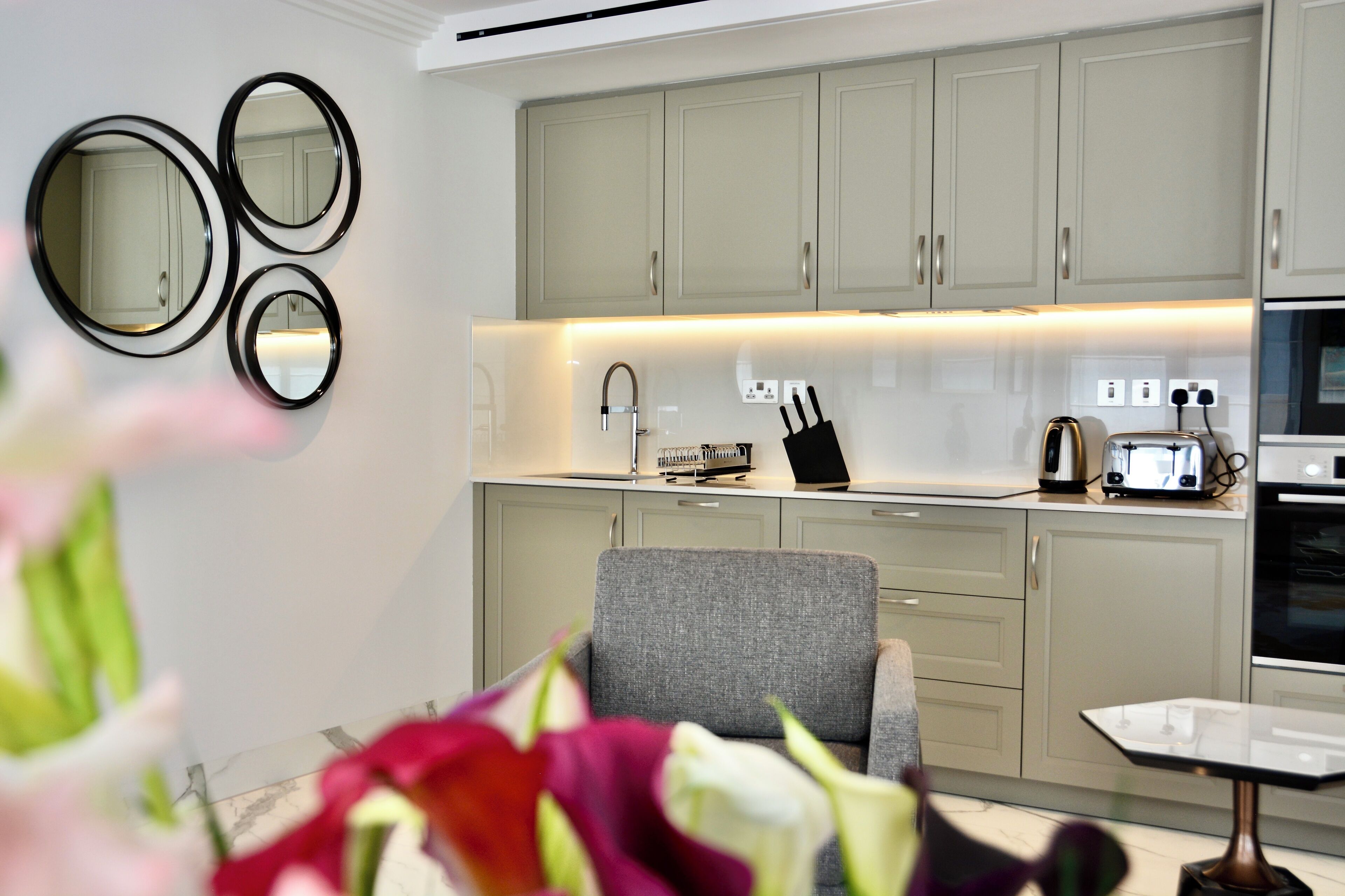 A modern kitchen with light-colored cabinets, a grey upholstered chair, a small table, and a countertop with a toaster and other appliances.