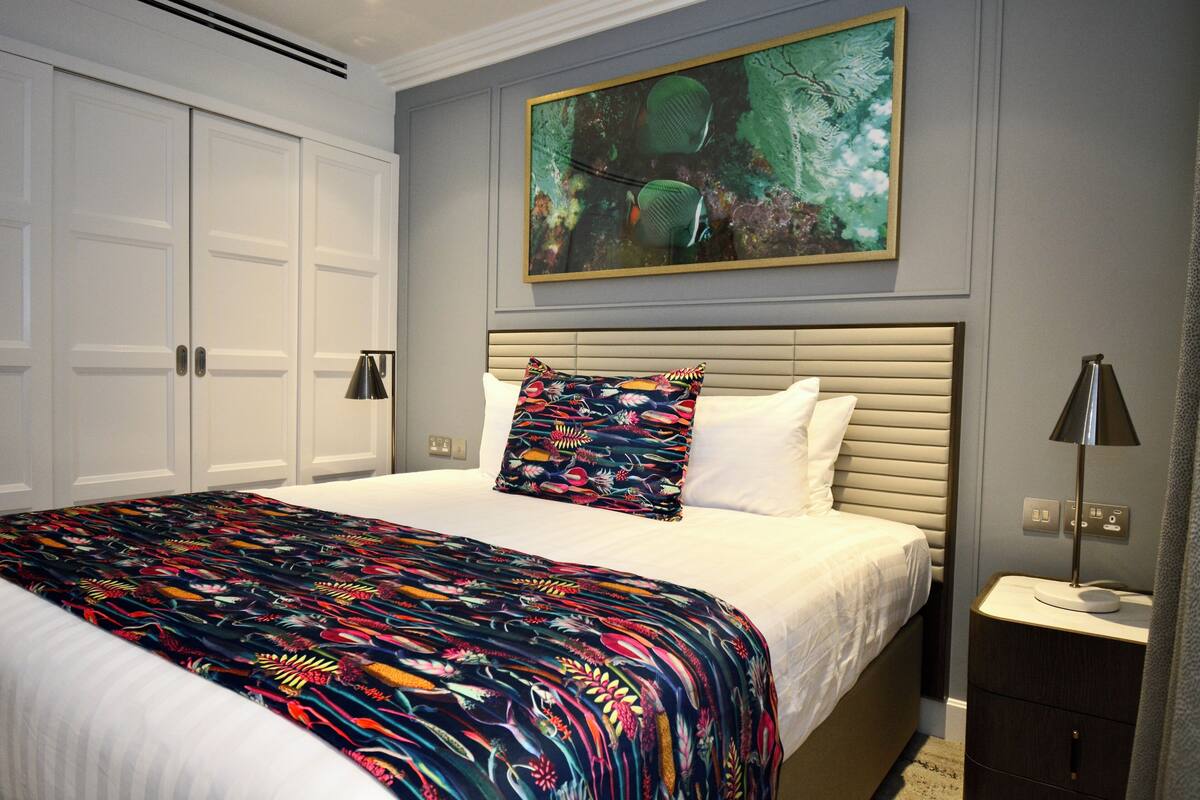 Een slaapkamer met een bed, een kleurrijk beddegoed, een hoofdbord, een nachtkastje, een lamp en een schilderij aan de muur.