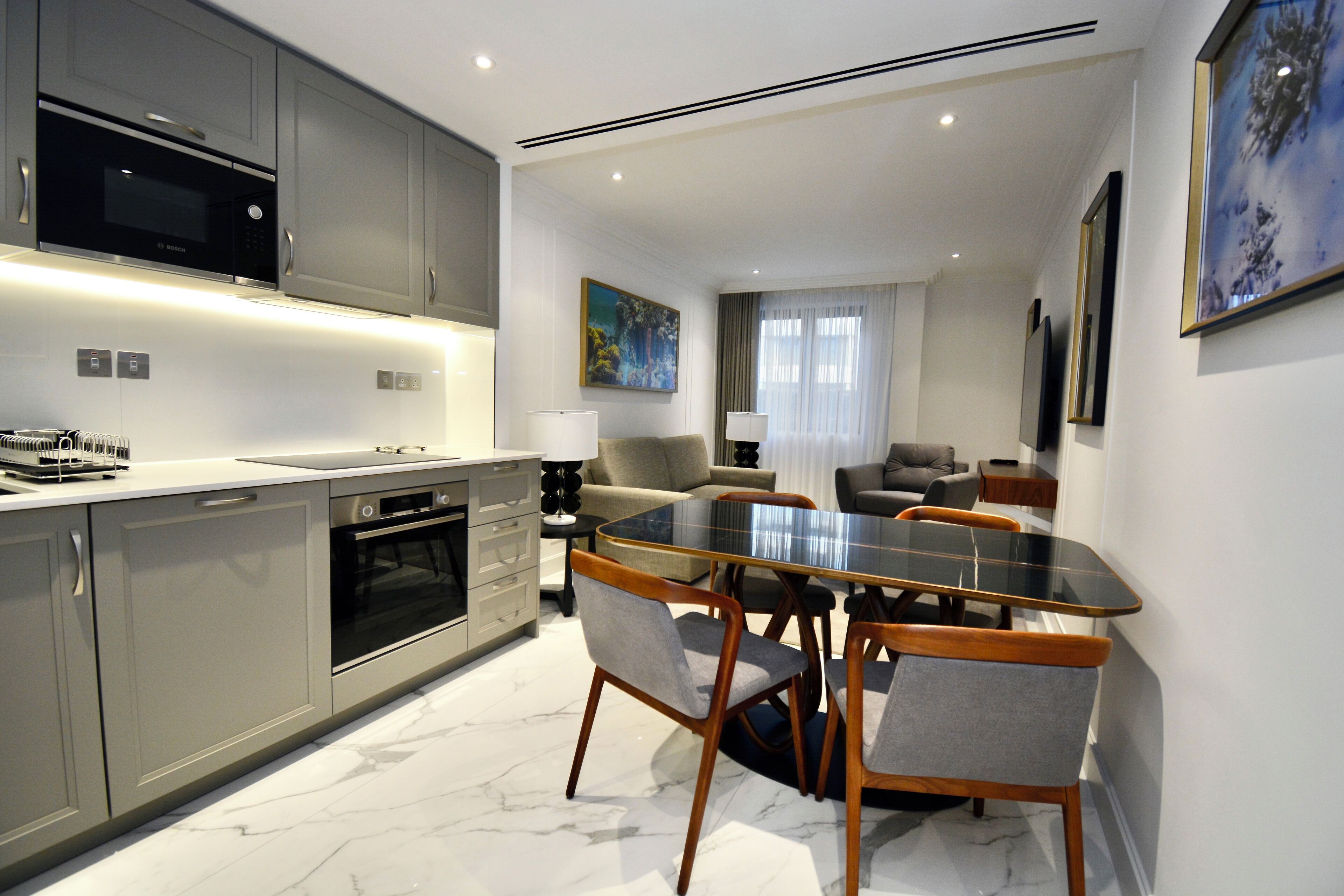 A modern kitchen with grey cabinets, a built-in oven, and a microwave. There is a dining area with a glass table and wooden chairs.