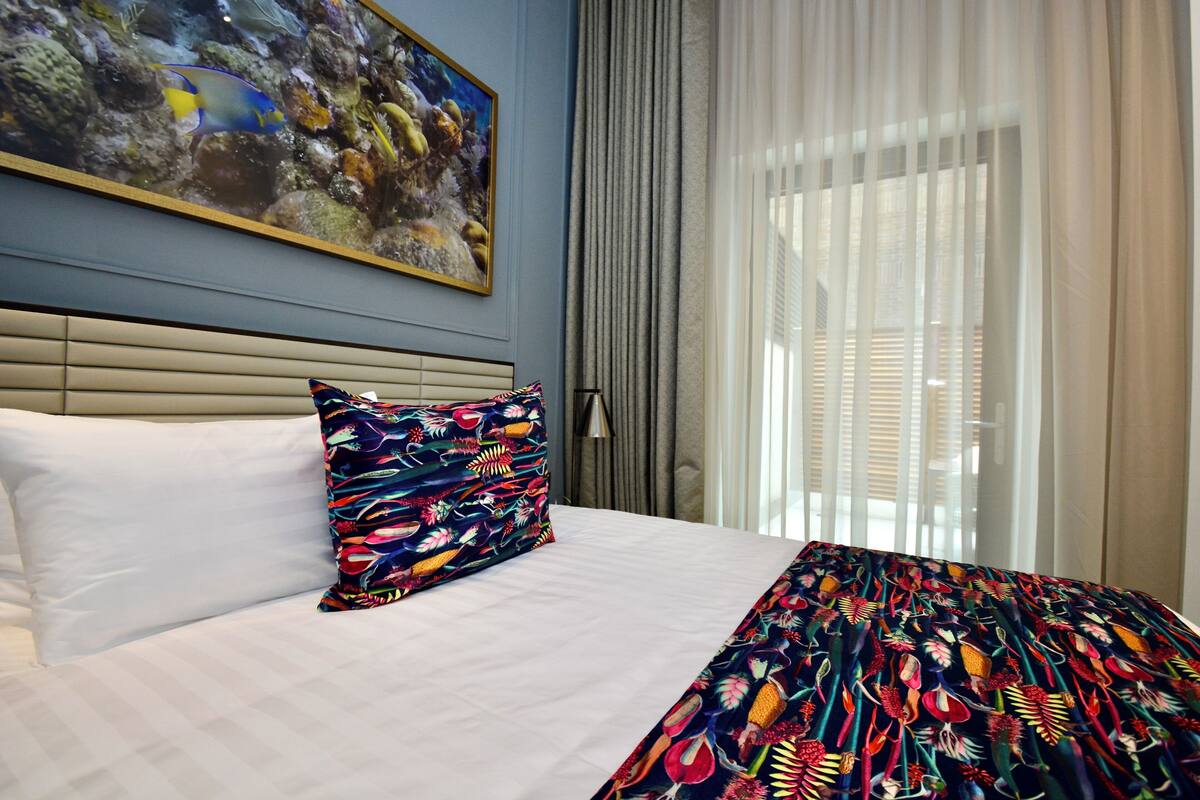 Een hotelkamer met een bed, een kleurrijk kussen, een schilderij van vissen aan de muur en dunne gordijnen.