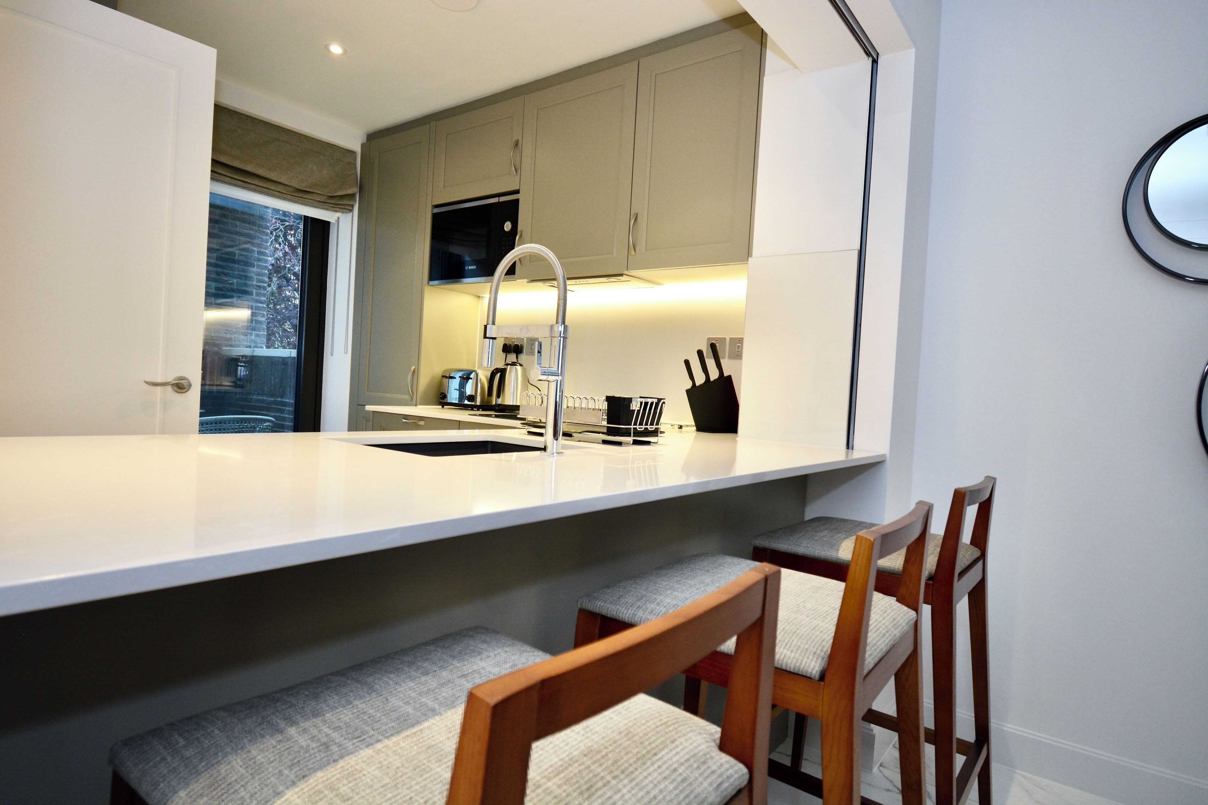 A modern kitchen with a white countertop, wooden chairs, and a built-in oven.