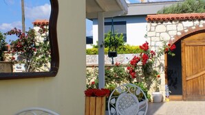 Une terrasse avec une table et des chaises en métal blanc, un banc en bois orné de fleurs rouges et un mur de pierre avec une porte en bois.