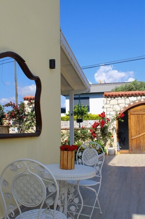 Une terrasse avec une table et des chaises en métal blanc, un banc en bois orné de fleurs rouges et un mur de pierre avec une porte en bois.