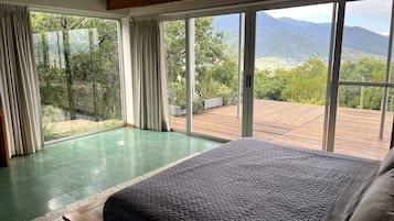 A bedroom with a large bed, a view of the outdoors through floor-to-ceiling windows, and a wooden deck outside.
