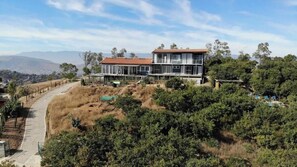 Une maison moderne à deux étages, dotée d’un toit plat, entourée d’une végétation luxuriante et baignée par un ciel dégagé.