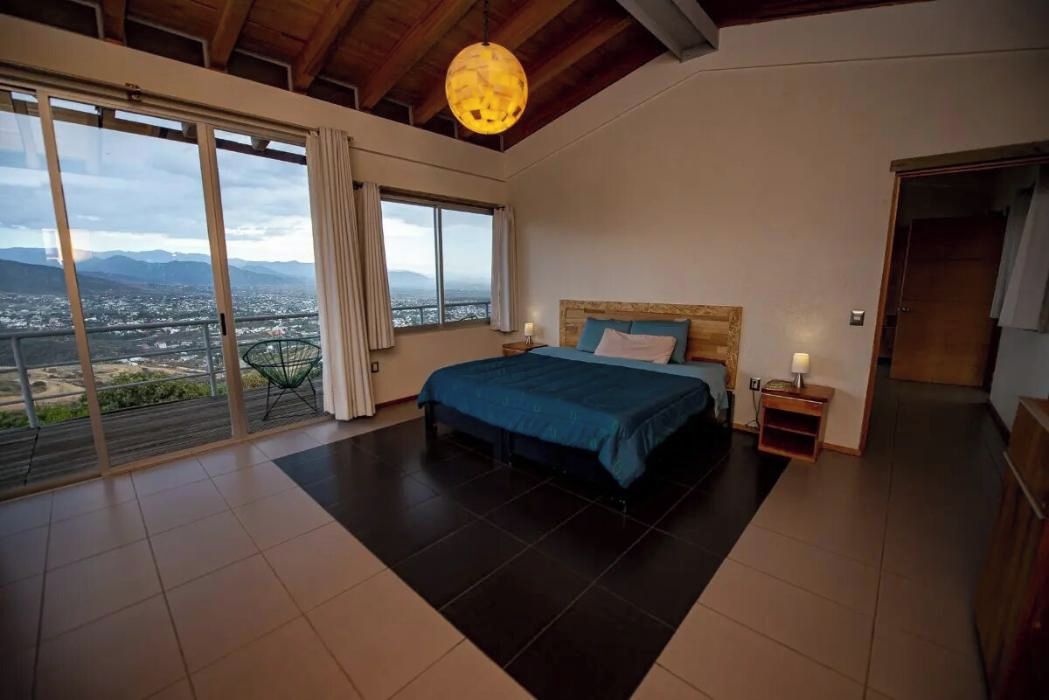 Ein Schlafzimmer mit einem großen Bett, einem Nachttisch, einer Lampe und Blick auf eine Stadtlandschaft durch eine Schiebetür.